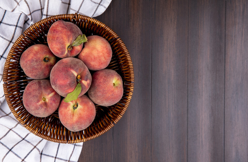 top view of fresh peaches on a bucket on a checked tablecloth on a wooden background with copy space
