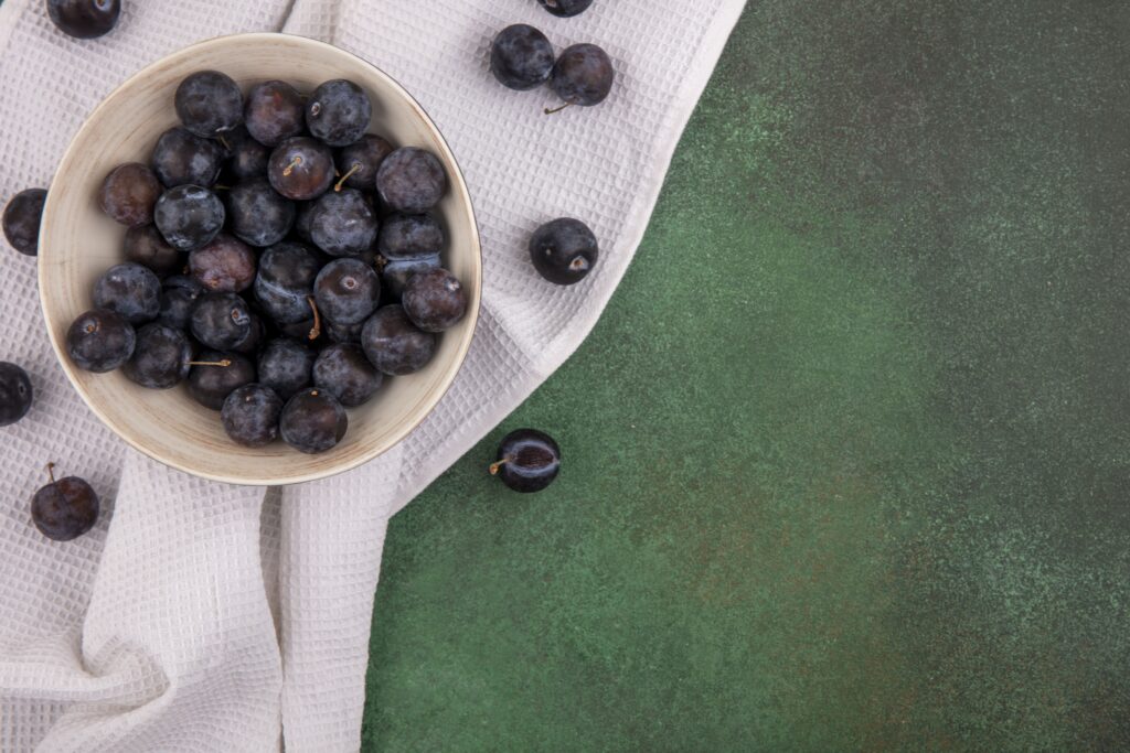 top view small sour blue black fruit sloes white bowl tablecloth green background with copy space (1)